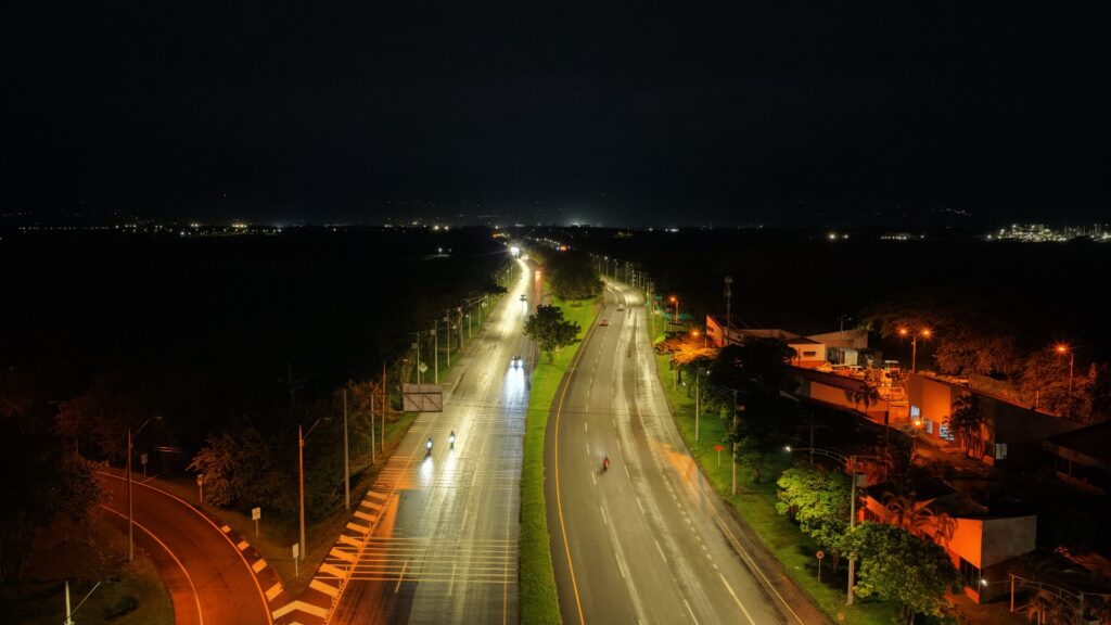 Fotografía aérea nocturna de una autopista de doble calzada con curvas suaves. La vía está intensamente iluminada por farolas de luz blanca, con varios vehículos y motocicletas dejando estelas luminosas a su paso. A la derecha, se observa un complejo de edificios industriales o comerciales con luces anaranjadas y zonas verdes. Al fondo, la oscuridad del paisaje rural se mezcla con pequeñas luces distantes bajo un cielo negro.