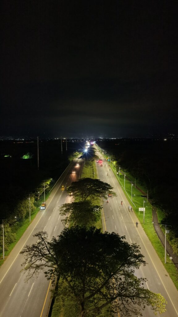 Fotografía aérea nocturna de la autopista de doble calzada iluminada Cali - Palmira. La carretera se extiende verticalmente hacia el horizonte, con postes de luz blanca a los lados y luces de vehículos en movimiento creando estelas brillantes. Grandes árboles se alinean en el separador central y en las orillas. Al fondo, se observan las luces distantes de una ciudad bajo un cielo oscuro.