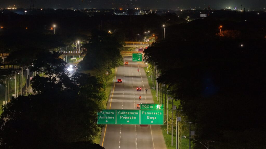 Fotografía aérea nocturna de una carretera de varios carriles. En primer plano, se observa un gran panel de señalización vial de color verde sobre la vía con indicaciones hacia destinos como Palmira, Candelaria, Popayán y Buga. Varios vehículos con sus luces encendidas circulan por la carretera, rodeada de árboles oscuros y postes de iluminación. Al fondo, se ven las luces de una zona urbana bajo un cielo nocturno despejado.